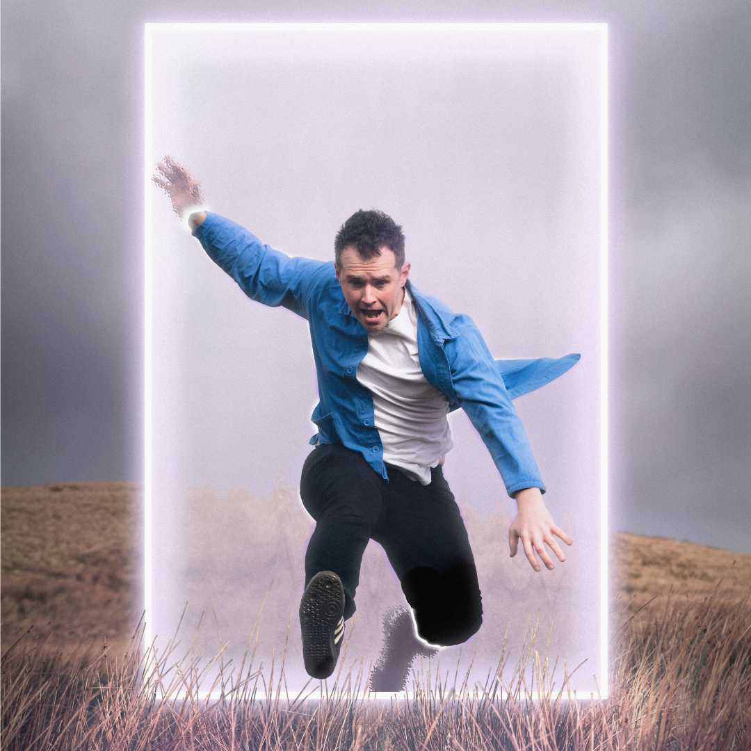 Òran - a male is jumping through a portal outside in a field. He's wearing a shirt, t-shirt and jeans.